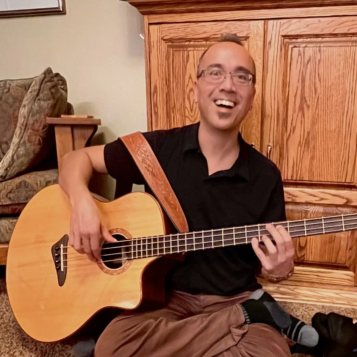 Jason Krivo Flores sitting on the floor at home, laughing, holding an acoustic bass guitar