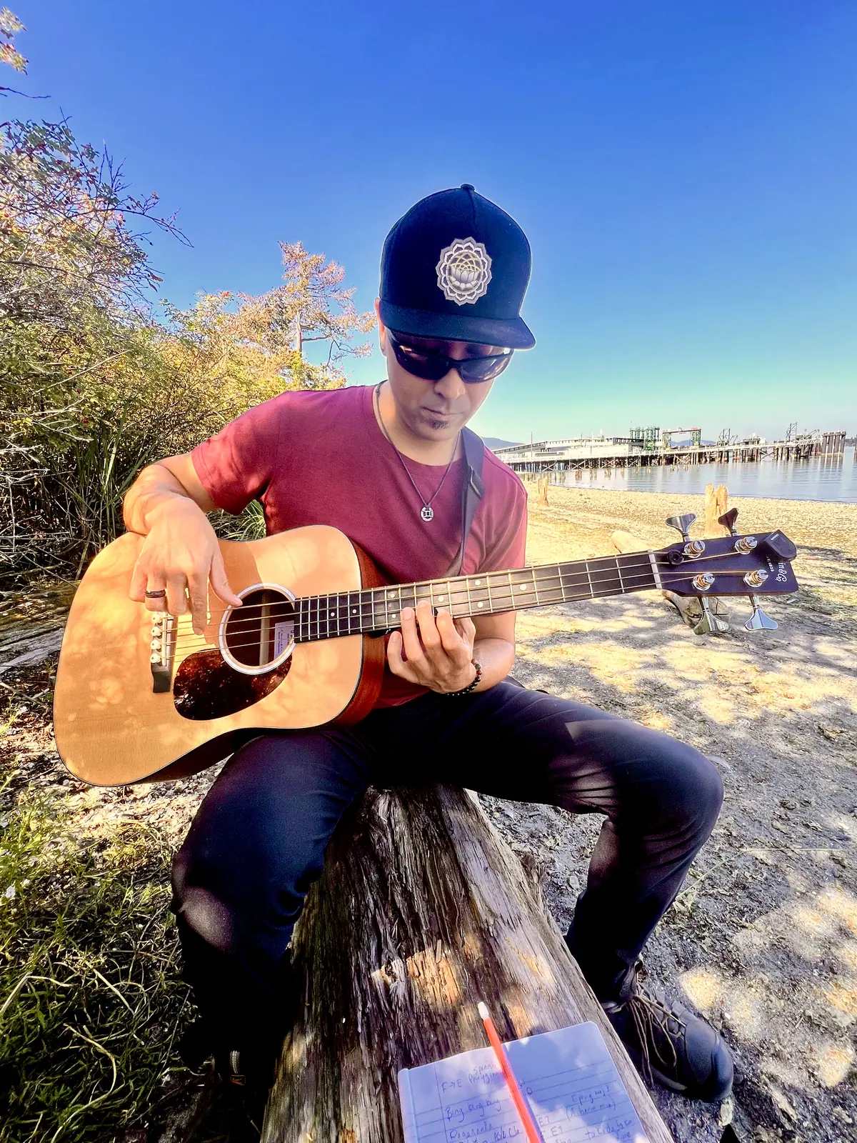 Playing a Martin Dreadnought Junior Bass seated on a driftwood log on a beach, a notebook of chord changes in the foreground