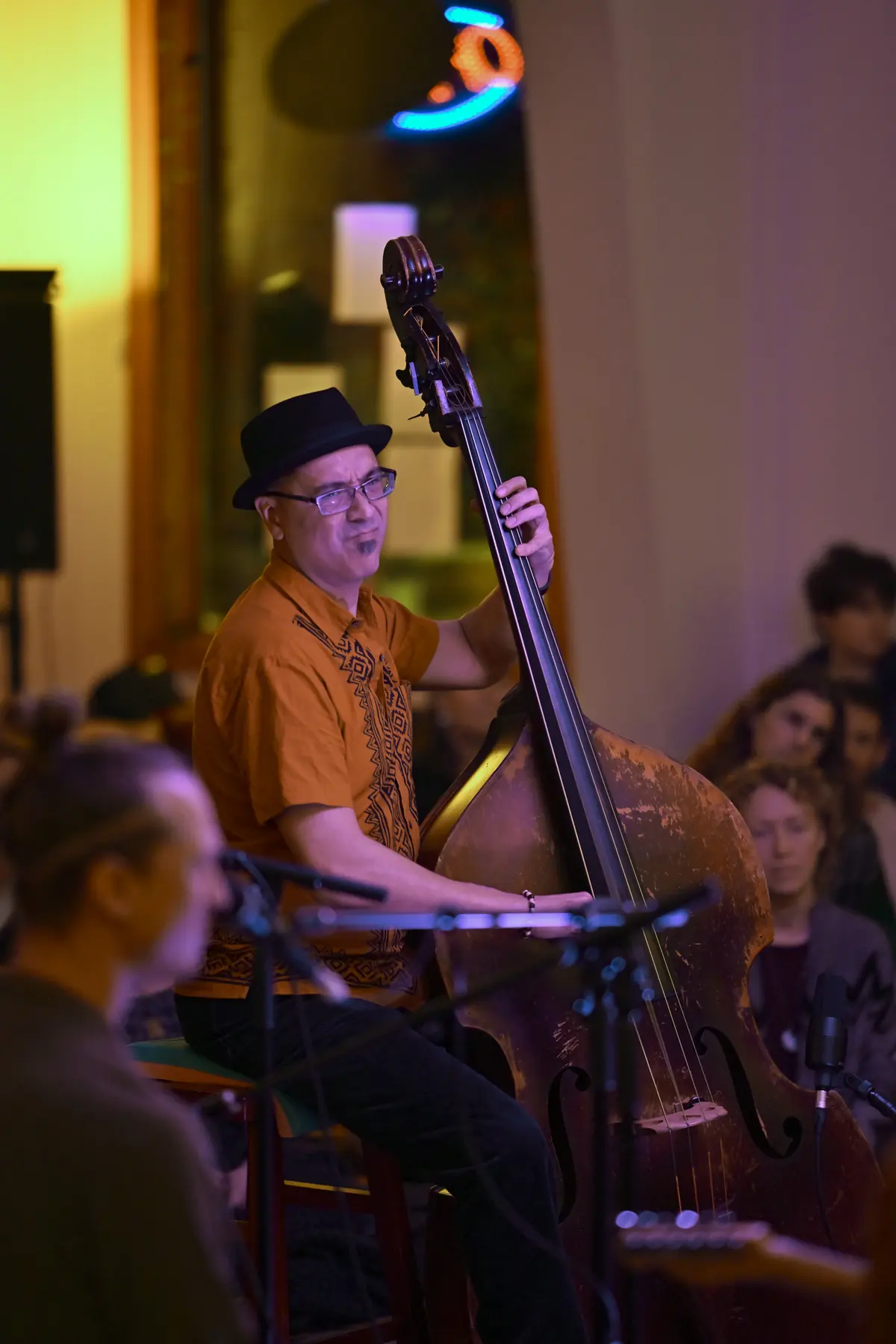 Jason Krivo Flores playing a weathered upright bass at a live show, seated, wearing a black hat